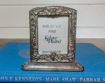 May include: A silver-plated photo frame with ornate detailing. The frame is rectangular with a decorative border and a small base. The text on the frame reads "SIZE: 2¼" x 3", FINE, Silver Plated, GODIERR".