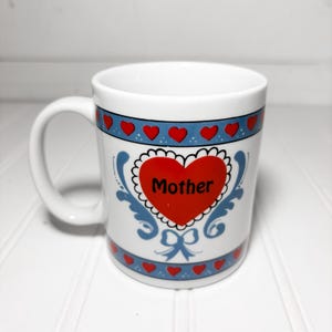 May include: White ceramic mug with a large red heart that says "Mother" in black text. The mug has a blue band with red hearts and decorative blue flourishes. A white handle is on the left side.