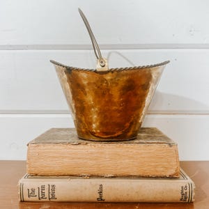 May include: A hammered copper bucket with a brass handle sits atop two antique books. The bucket has a textured surface and a decorative rope-like trim around the rim. The books are aged with visible wear and tear.