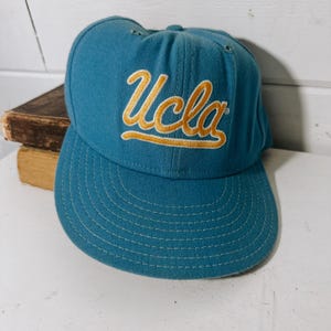 May include: A blue and gold UCLA baseball cap. The cap has a curved brim and a stitched design.