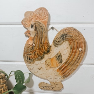 May include: A ceramic rooster wall hanging with brown, orange, and white paint. The rooster has a detailed design and a hanging loop on the top.