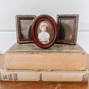 May include: Three antique picture frames with red and gold trim, one oval and two rectangular, sit atop two vintage books. The oval frame holds a sepia-toned portrait of a young girl. The books have aged, cream-colored pages.
