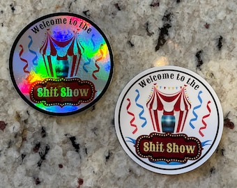 May include: Two round stickers with a holographic and white background. Both stickers feature a red and white striped tent with a blue top and the text "Welcome to the Shit Show".