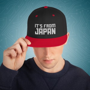 May include: A black and red baseball cap with the text "IT'S FROM JAPAN" in white lettering.