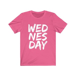 Puede incluir: Camiseta rosa de manga corta con el texto "WEDNES DAY" en blanco, con letras estilizadas.