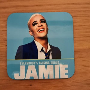 May include: A blue square coaster with a picture of a person wearing a blue suit and a white shirt with a blue and yellow tie. The text "EVERYBODY'S TALKING ABOUT JAMIE" is printed on the coaster.