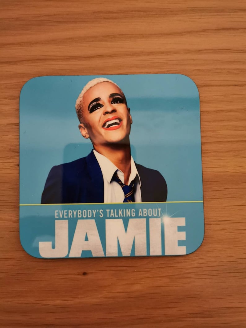 May include: A blue square coaster with a picture of a person wearing a blue suit and a white shirt with a blue and yellow tie. The text "EVERYBODY'S TALKING ABOUT JAMIE" is printed on the coaster.