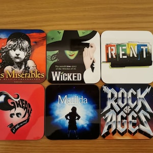 May include: Six square coasters featuring the logos of popular Broadway musicals. The coasters are white with colorful images and text. The musicals featured are Les Miserables, Wicked, Rent, Grease, Matilda, and Rock of Ages.