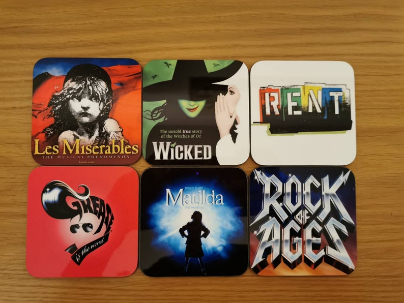 May include: Six square coasters featuring the logos of popular Broadway musicals. The coasters are white with colorful images and text. The musicals featured are Les Miserables, Wicked, Rent, Grease, Matilda, and Rock of Ages.