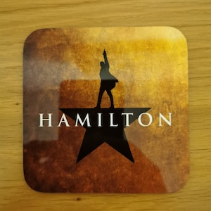 May include: A brown and gold coaster with a black silhouette of a person standing on a black star. The text "HAMILTON" is printed in white below the star.