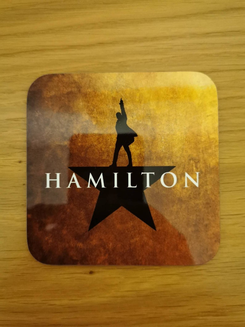 May include: A brown and gold coaster with a black silhouette of a person standing on a black star. The text "HAMILTON" is printed in white below the star.
