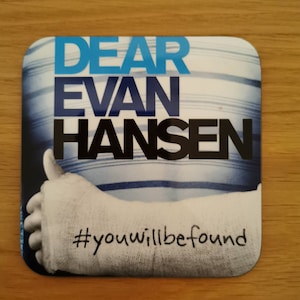 May include: A white coaster with blue stripes and the text "DEAR EVAN HANSEN #youwillbefound". The coaster features a photo of a person's arm in a cast giving a thumbs up.