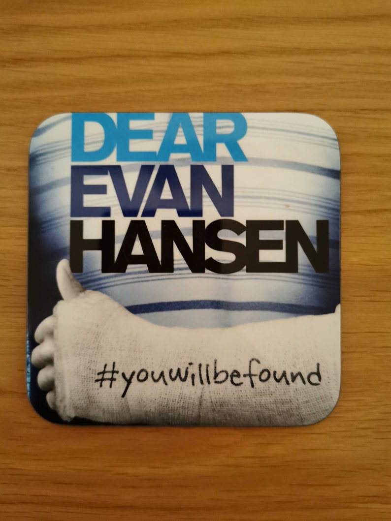 May include: A white coaster with blue stripes and the text "DEAR EVAN HANSEN #youwillbefound". The coaster features a photo of a person's arm in a cast giving a thumbs up.