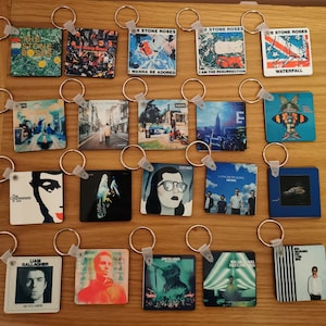 May include: A collection of 16 keychains featuring album covers from various artists, including The Stone Roses, Liam Gallagher, and Courteeners. The keychains are arranged in a grid pattern on a wooden surface.