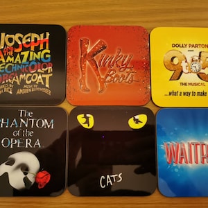 May include: Six square coasters featuring various Broadway show logos. The coasters are black, red, yellow, blue, and white with colorful text and images. The shows featured are Joseph and the Amazing Technicolor Dreamcoat, Kinky Boots, 9 to 5, The Phantom of the Opera, Cats, and Waitress.