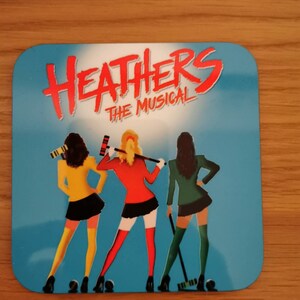 May include: A blue and white square coaster with the text "Heathers The Musical" in red and white. Three women in different colored outfits are holding lacrosse sticks.