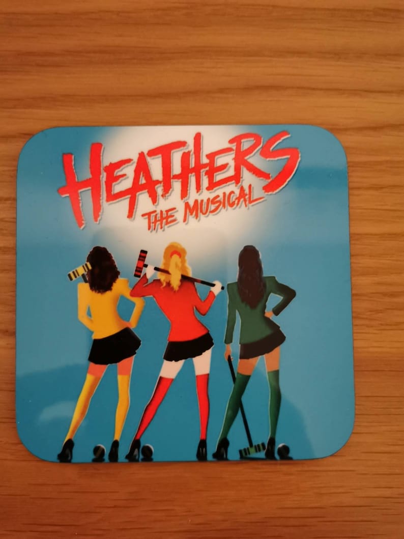 May include: A blue and white square coaster with the text "Heathers The Musical" in red and white. Three women in different colored outfits are holding lacrosse sticks.
