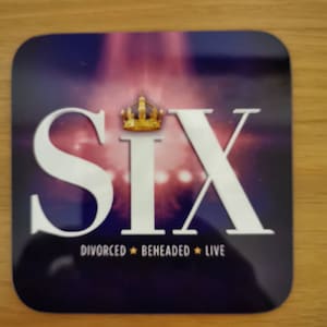 May include: A purple and white coaster with the word "SIX" in large white letters and a gold crown above the word. The text "DIVORCED  BEHEADED  LIVE" is below the word "SIX".