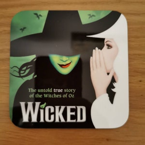 May include: A square, black and white coaster with a green and black image of two witches from the musical Wicked. The text "The untold true story of the Witches of Oz" and "WICKED" are printed on the coaster.