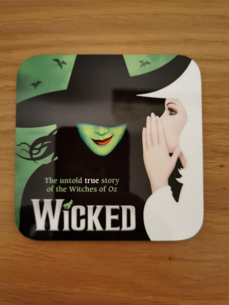 May include: A square, black and white coaster with a green and black image of two witches from the musical Wicked. The text "The untold true story of the Witches of Oz" and "WICKED" are printed on the coaster.