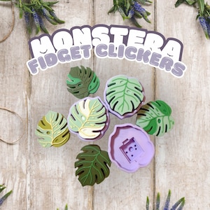 May include: A collection of Monstera Fidget Clickers in various shades of green and purple, shaped like Monstera leaves. The text "MONSTERA FIDGET CLICKERS" is displayed above the fidget toys. The toys are arranged on a light wooden surface.