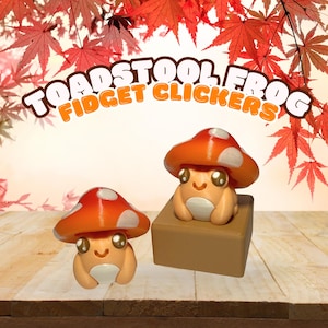 May include: Two orange and white toadstool frog fidget clickers. The frogs have smiling faces and are sitting on a brown surface. The text "TOADSTOOL FROG FIDGET CLICKERS" is arched above the frogs.