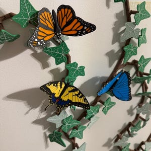 May include: A decorative vine with green ivy leaves and brown branches, adorned with three colorful butterflies. The butterflies include a monarch with orange and black wings, a yellow and black swallowtail, and a blue butterfly.