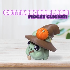 May include: A green frog-shaped fidget clicker wearing a brown witch hat with an orange pumpkin on top. The text "COTTAGECORE FROG FIDGET CLICKER" is above the frog. The frog has large black eyes and a small brown stem in its mouth.
