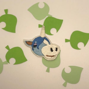 Peut inclure: Un patch brodé représentant un personnage animal bleu et blanc avec une corne. Le personnage a un visage blanc avec des yeux noirs et un nez rose. Le patch est entouré de découpes en forme de feuilles vertes.