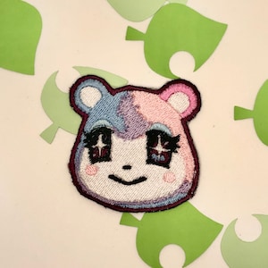 May include: Embroidered patch of a pink and blue animal character with sparkling eyes. The character has a friendly expression and is smiling.