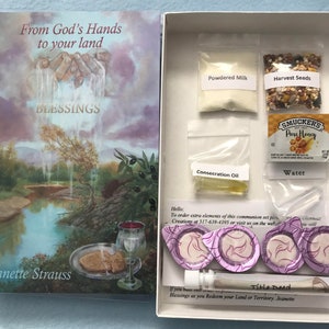 May include: A white box with a book titled "From God's Hands to your land BLESSINGS" by Jeanette Strauss. The box contains a communion set with items like powdered milk, harvest seeds, honey, water, consecration oil, and wafers.