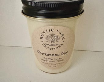 May include: A soy wax candle in a glass jar with a black metal lid. The label reads Rustic Farm Creations, Christmas Day, Handmade in Florida, and 6oz | 170g. The candle is off-white and features a graphic of a small house with sun rays.