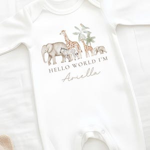 Pode incluir: Macacão branco para bebé com mangas compridas, com uma ilustração em aquarela de um elefante, uma girafa, uma zebra e outros animais. O texto diz "HELLO WORLD I'M Ariella". O macacão tem fechos de pressão.
