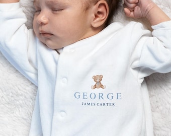 Könnte beinhalten: Ein schlafendes Baby in einem weißen Strampler mit einem Teddybären-Motiv und dem Namen "GEORGE JAMES CARTER" in blauer Schrift. Das Baby liegt auf einer flauschigen, weißen Decke.