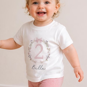 May include: White t-shirt with a floral wreath design, a pink bow, and the number 2. The name "Bella" is written below the number. The shirt is worn by a toddler wearing pink leggings.