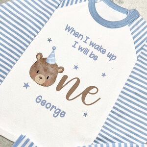May include: A baby's first birthday pyjama set. The white top features a brown bear with a party hat and the text "When I wake up I will be one George". The sleeves and trousers are blue and white striped.