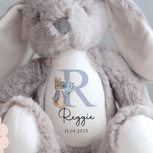 Peut inclure: Une peluche lapin grise avec de longues oreilles et des accents blancs sur les pattes et l'int&eacute;rieur des oreilles. Le lapin porte une inscription personnalis&eacute;e sur sa poitrine avec le nom "Reggie" et la date 11.04.2025, ainsi qu'un "R" bleu et une illustration de Pierre Lapin.