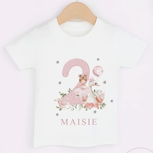 Pu&ograve; includere: T-shirt bianca con un disegno rosa del numero 2 con una principessa, un cigno ed elementi floreali. Il nome "Maisie" &egrave; stampato sotto il disegno. La maglietta &egrave; appesa a un appendiabiti di legno.