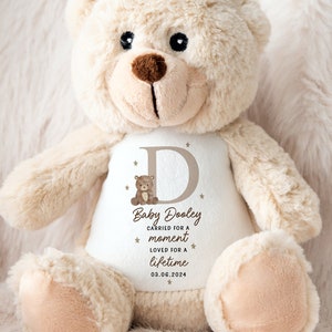 Könnte beinhalten: Ein beiger Teddybär mit einem weißen Shirt mit dem Namen "Baby Dooley", einem Buchstaben "D", einer kleinen Teddybär-Grafik und dem Text "carried for a moment, loved for a lifetime, 03.06.2024". Der Bär hat eine braune Nase und schwarze Augen.