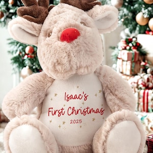 May include: A plush reindeer toy with brown antlers, a red nose, and a cream-colored body. The text "Isaac's First Christmas 2025" is printed on the reindeer's chest. The background includes a Christmas tree and wrapped gifts.