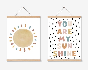 May include: Two framed art prints. One features a watercolor sun in shades of yellow, orange, and blue. The other print has the words "YOU ARE MY SUNSHINE" in colorful letters against a white background with black polka dots. Both are hung with wooden bars.