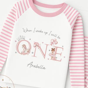 以下が含まれることがあります： 「When I wake up I will be ONE Arabella」の文字が入った、白とピンクのストライプのパジャマセット。長袖トップスはピンクの袖とピンクのネックライン。デザインにはユニコーン、バレリーナ、風船が含まれています。