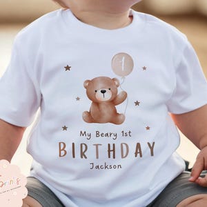 Może przedstawiać: Biała koszulka z brązowym misiem trzymającym balon z cyfrą 1. Pod misiem wydrukowano napis "My Beary 1st BIRTHDAY Jackson". Małe brązowe gwiazdki rozrzucone są wokół wzoru.