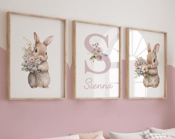 Könnte beinhalten: Drei gerahmte Kunstdrucke. Zwei zeigen ein braunes Kaninchen mit einem Blumenstrauß. Der dritte Druck hat einen großen Buchstaben "S" mit floralen Akzenten und dem Namen "Sienna" in einem zarten Rosa. Die Rahmen sind aus hellem Holz.
