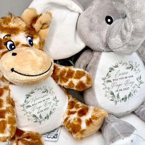Puede incluir: Dos peluches, una jirafa y un elefante. La jirafa es marrón y blanca, el elefante es gris. Ambos tienen una camiseta blanca con el texto "Will you be our Page Boy" y un nombre y una fecha. La camiseta de la jirafa dice "Harrison 19.06.2027". La camiseta del elefante dice "Lucas 19.06.2027".
