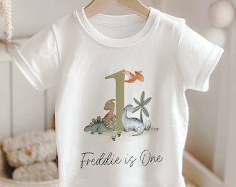 May include: White short-sleeved t-shirt featuring a dinosaur-themed design. The design includes the number "1", several dinosaurs, and the text "Freddie is One". The shirt is hanging on a wooden hanger.