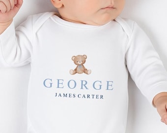 Könnte beinhalten: Weißer Baby-Strampler mit langen Ärmeln und Füßen. Der Strampler hat eine Aquarell-Teddybär-Grafik über dem Namen "GEORGE" in Blau, mit "JAMES CARTER" darunter. Das Baby liegt auf einer weißen Oberfläche.
