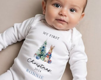 May include: A white baby onesie with long sleeves. The onesie features a watercolor illustration of a Christmas tree, a rabbit, and wrapped gifts. Text on the onesie reads "MY FIRST Christmas 2024" and a name.