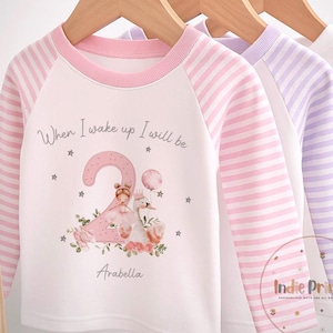 May include: Three long-sleeved shirts hanging on a wooden rack. The first shirt is white with pink striped sleeves and a pink neckline. It features a graphic with the text "When I wake up I will be 2 Arabella". The other shirts are pink and white striped and solid white.