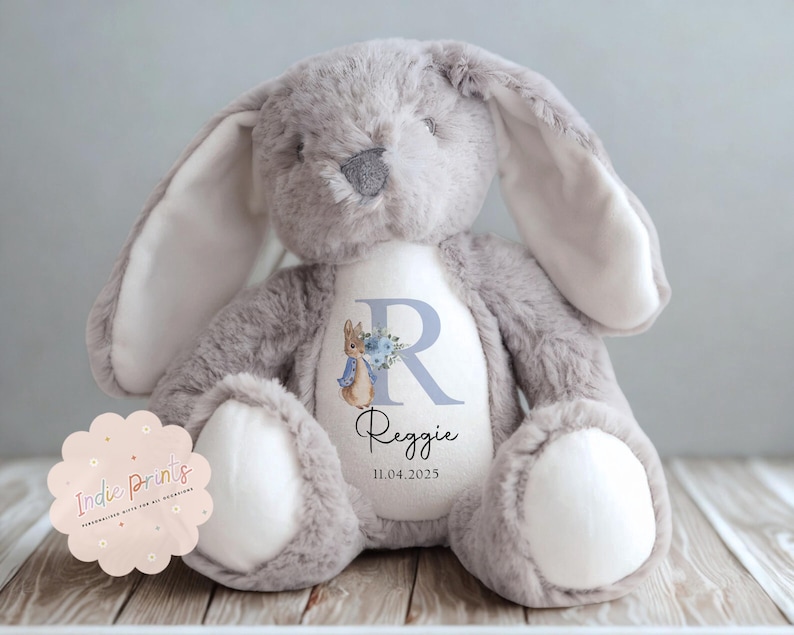 Peut inclure: Une peluche lapin grise avec de longues oreilles et des accents blancs sur les pattes et l'int&eacute;rieur des oreilles. Le lapin porte une inscription personnalis&eacute;e sur sa poitrine avec le nom "Reggie" et la date 11.04.2025, ainsi qu'un "R" bleu et une illustration de Pierre Lapin.